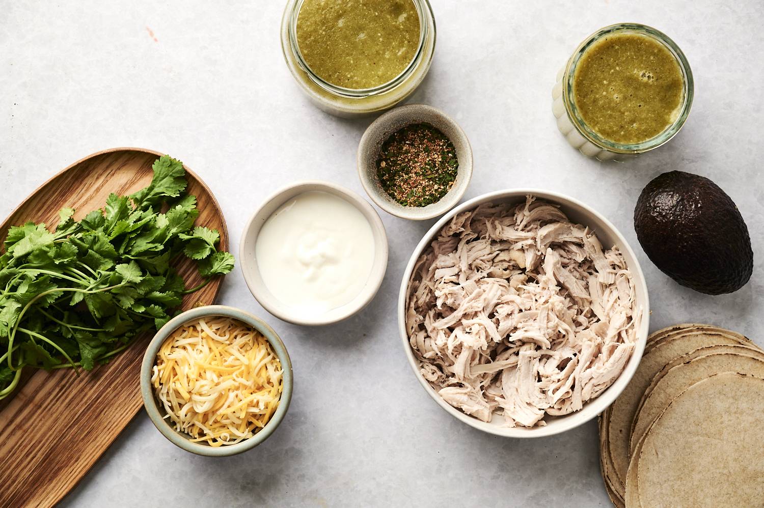 Ingredients for green chicken enchiladas, including shredded chicken, tortillas, shredded cheese, cilantro, sour cream, avocado, and green enchilada sauce.