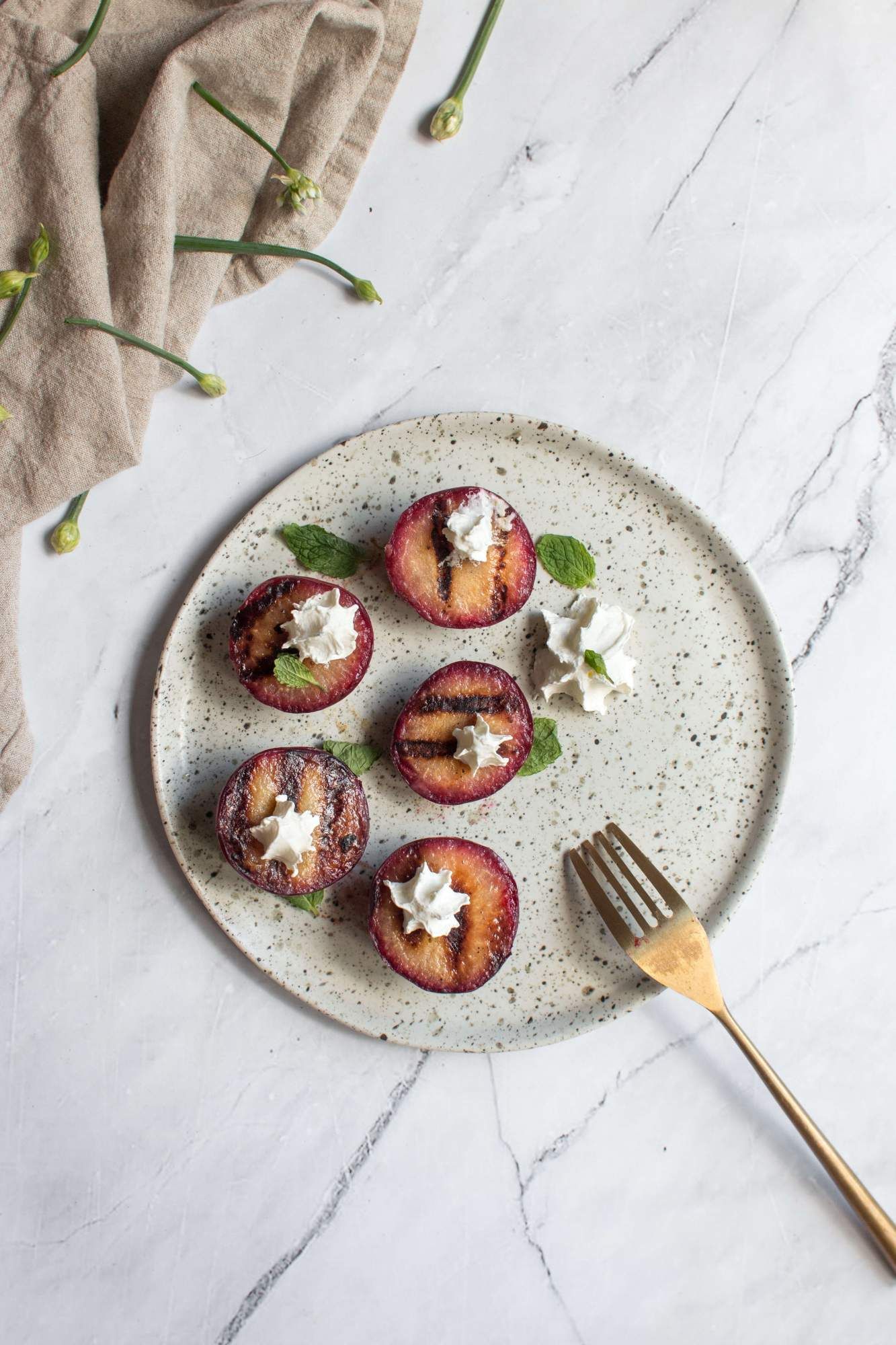 Plums that ave been grilled on a plate with fresh herbs, whipped cream, and cinnamon.