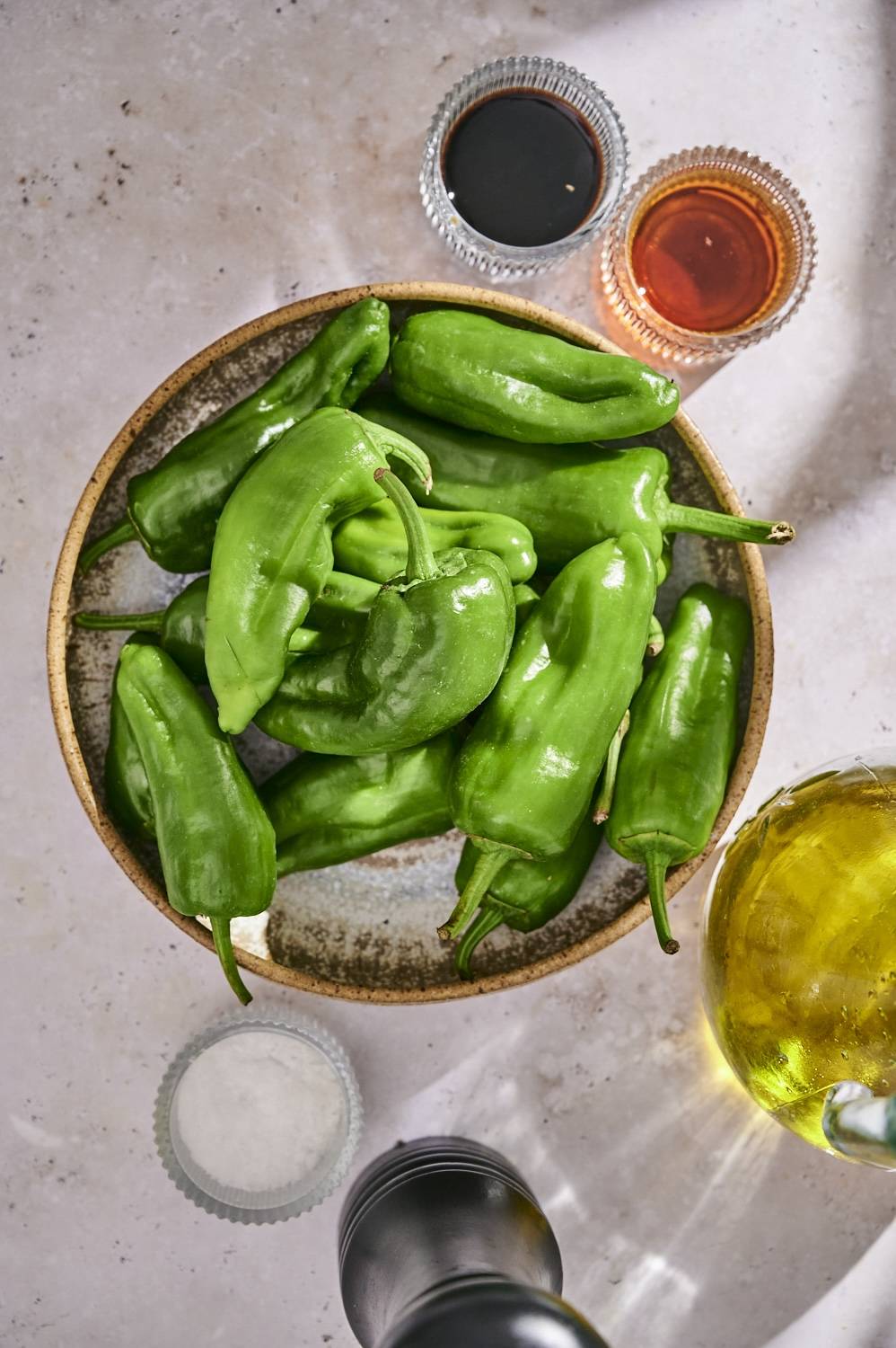 Shishito peppers in a bowl with sesame oil, soy sauce, olive oil, salt, and pepper on the side.