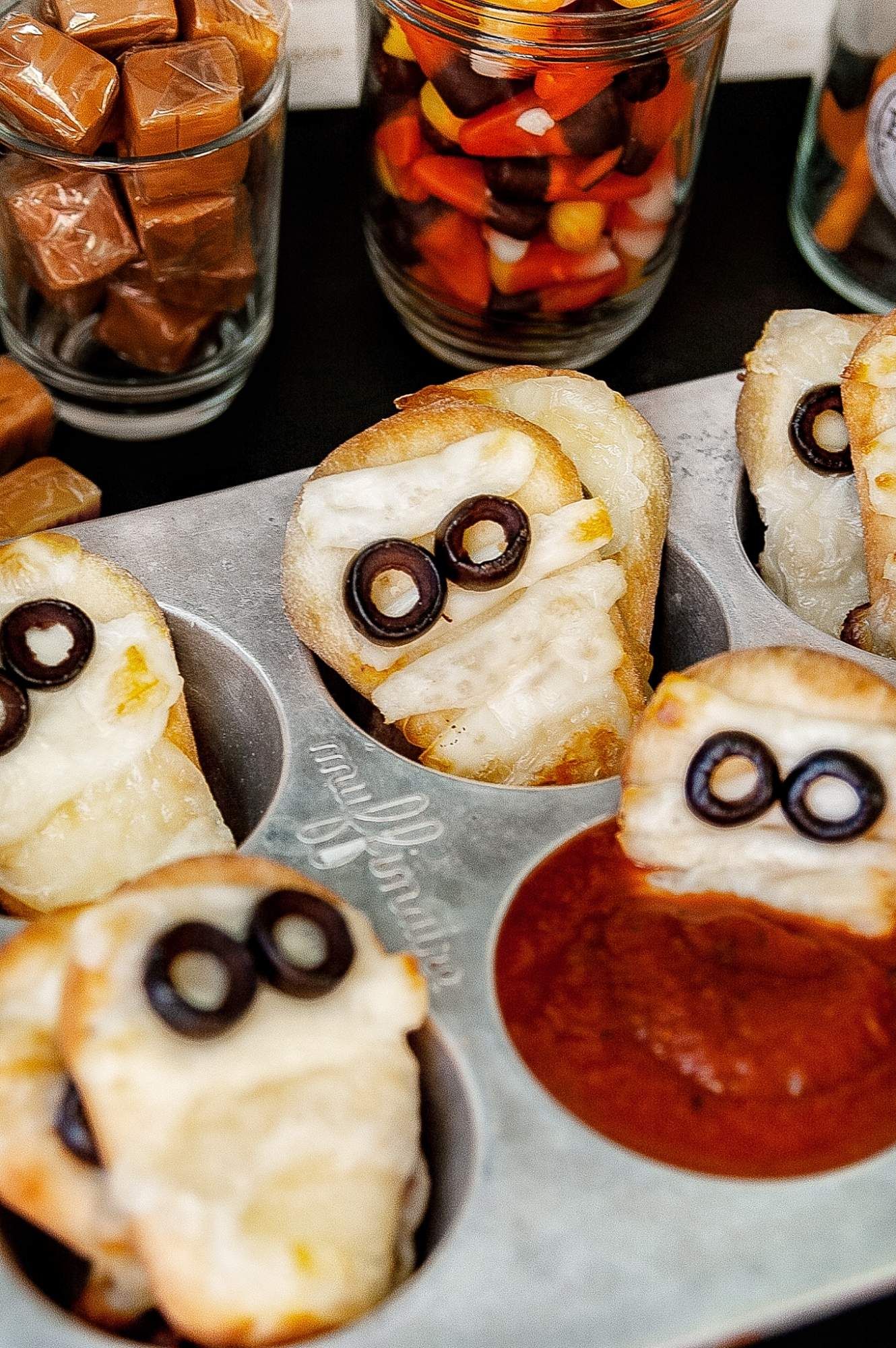 Pizza dippers that look like mummies in a muffin tin with candy in the background.