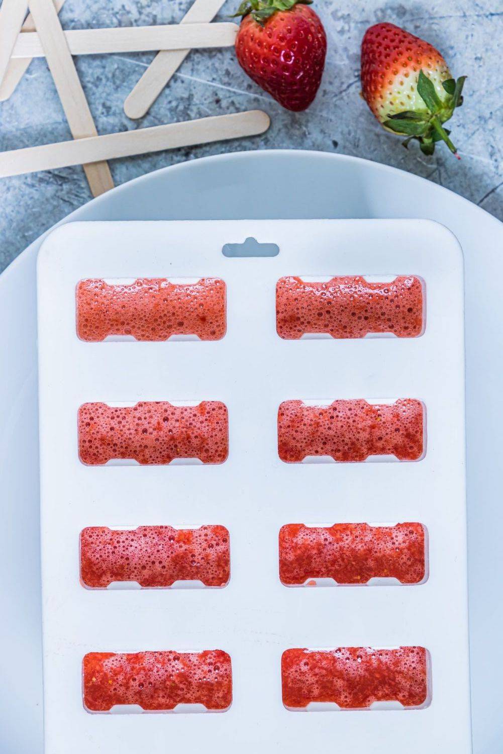 Strawberry popsicle mix being poured into a plastic mold with wooden popsicle sticks on the side.