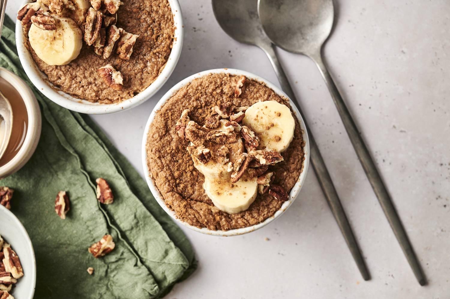 Overhead view of two ramekins filled with baked protein oats, topped with banana slices, pecans, and a sprinkle of cinnamon. A green napkin and two spoons are placed alongside the ramekins, with scattered pecan pieces on the surface.