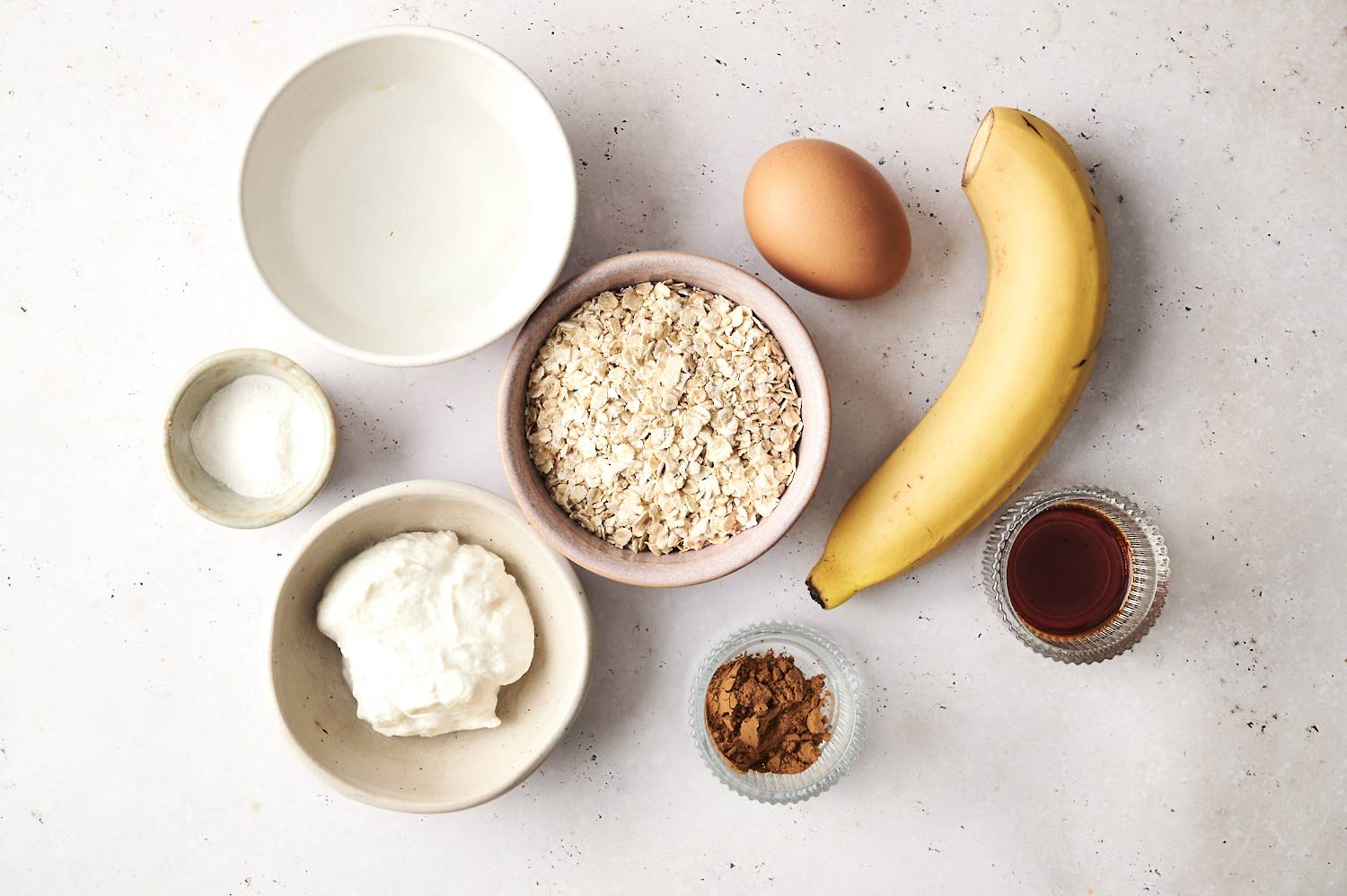  Ingredients for protein baked oats arranged on a light surface, including a banana, an egg, a bowl of rolled oats, a bowl of yogurt, a small dish of cocoa powder, a small dish of baking powder, a bowl of water, and a dish of vanilla extract.