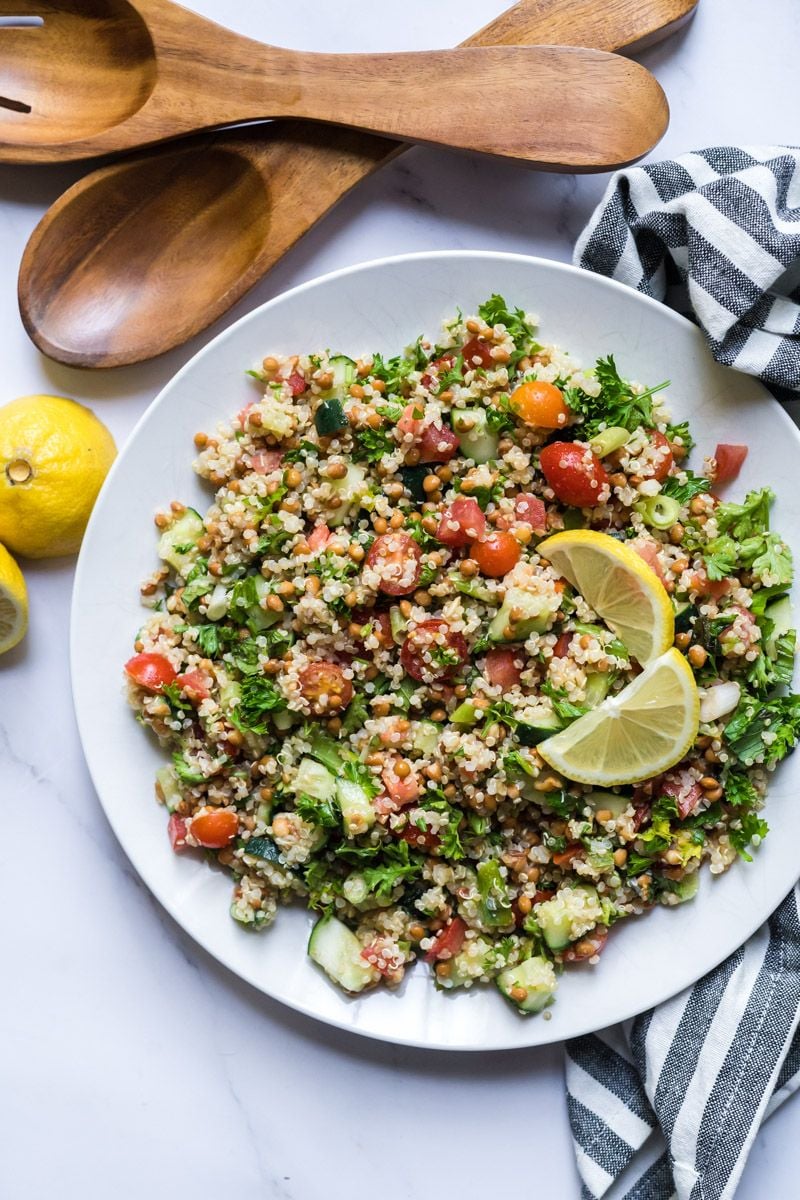 Tabbouleh made with quinoa, lentils, tomatoes, cucumbers, parsley, and mint on a plate with wooden utensils.