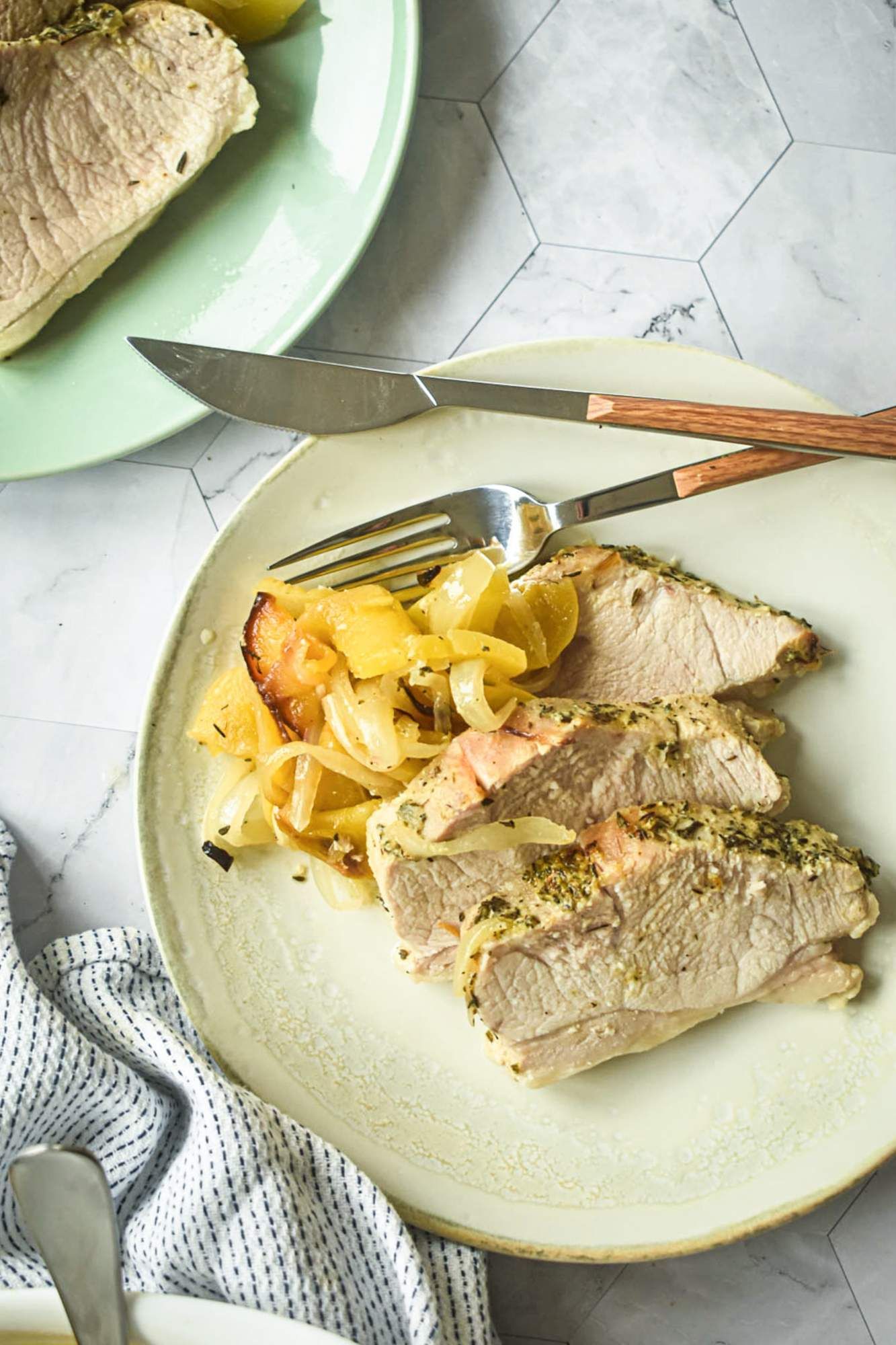 Roasted pork coated in herbs with slices apples and onions on a plate with a knife and fork.