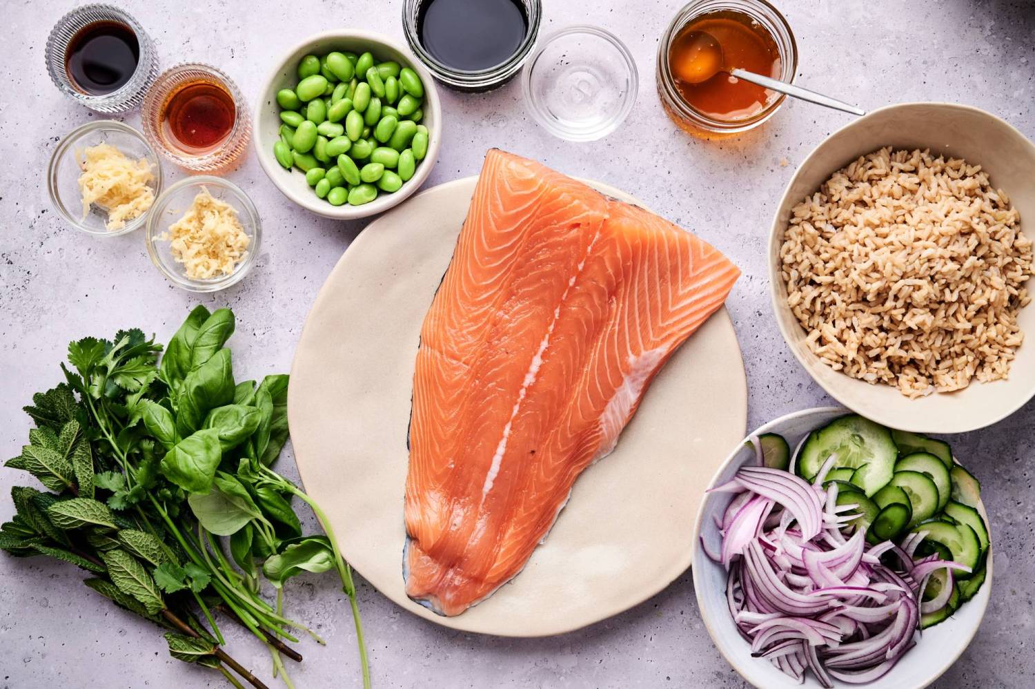 Ingredients for salmon rice bowls including raw salmon, brown rice, mint, cilantro, basil, edamame, cucumber, red onion, soy sauce, and honey.