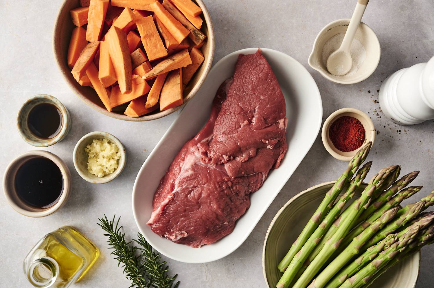 An overhead view of fresh ingredients for a sheet pan meal: raw steak, sliced sweet potatoes, fresh asparagus, minced garlic, olive oil, seasonings, and a sprig of rosemary.