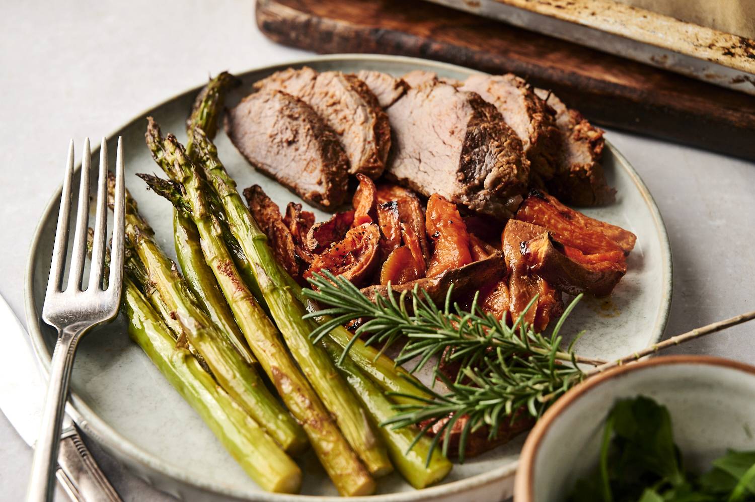 A beautifully plated dish showcasing tender rosemary-seasoned steak slices, roasted asparagus spears, and caramelized sweet potato wedges, garnished with a sprig of fresh rosemary.