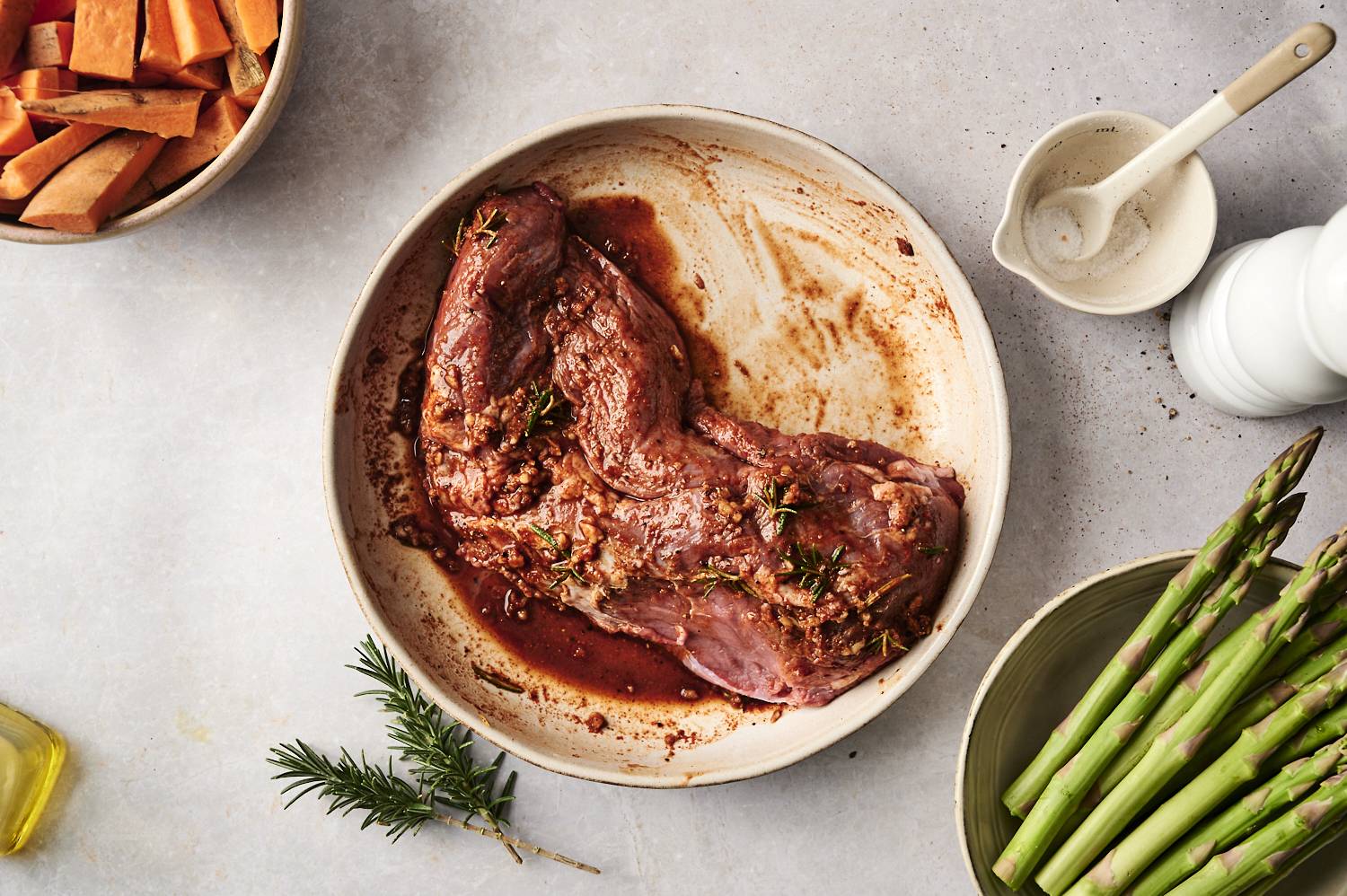 An overhead view of fresh ingredients for a sheet pan meal: raw steak, sliced sweet potatoes, fresh asparagus, minced garlic, olive oil, seasonings, and a sprig of rosemary