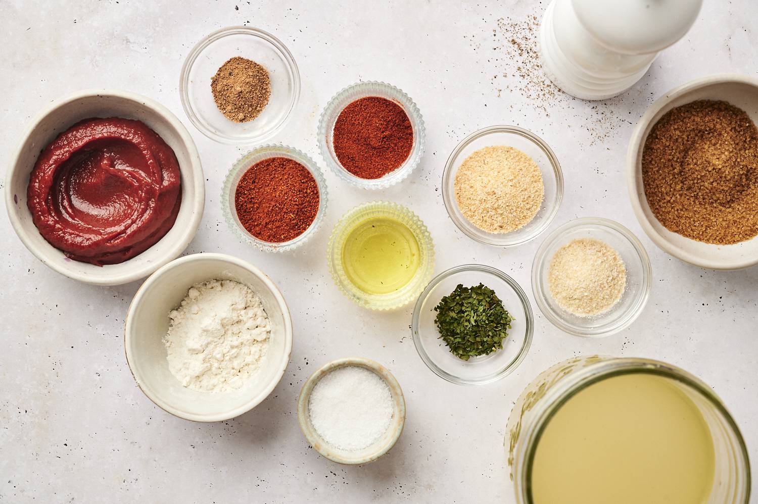 Flat lay view of ingredients for homemade enchilada sauce including tomato paste, flour, chili powder, garlic powder, onion powder, cumin, brown sugar, oil, broth, salt and dried herbs arranged in small bowls.