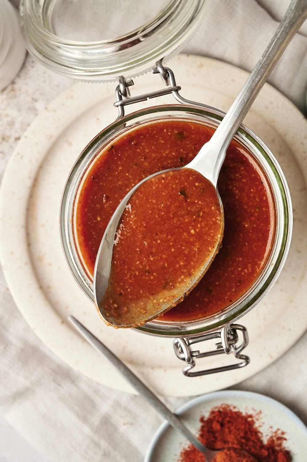 Close-up of a glass jar filled with homemade enchilada sauce, a portion lifted with a spoon to show its smooth texture. The jar is placed on a ceramic plate with a small dish of paprika next to it.