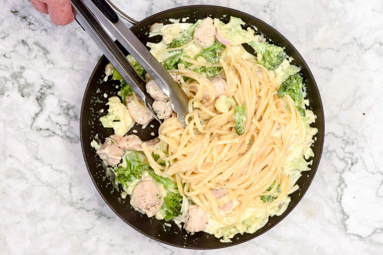 Healthy chicken broccoli alfredo in a skillet with tongs stirring the pasta.