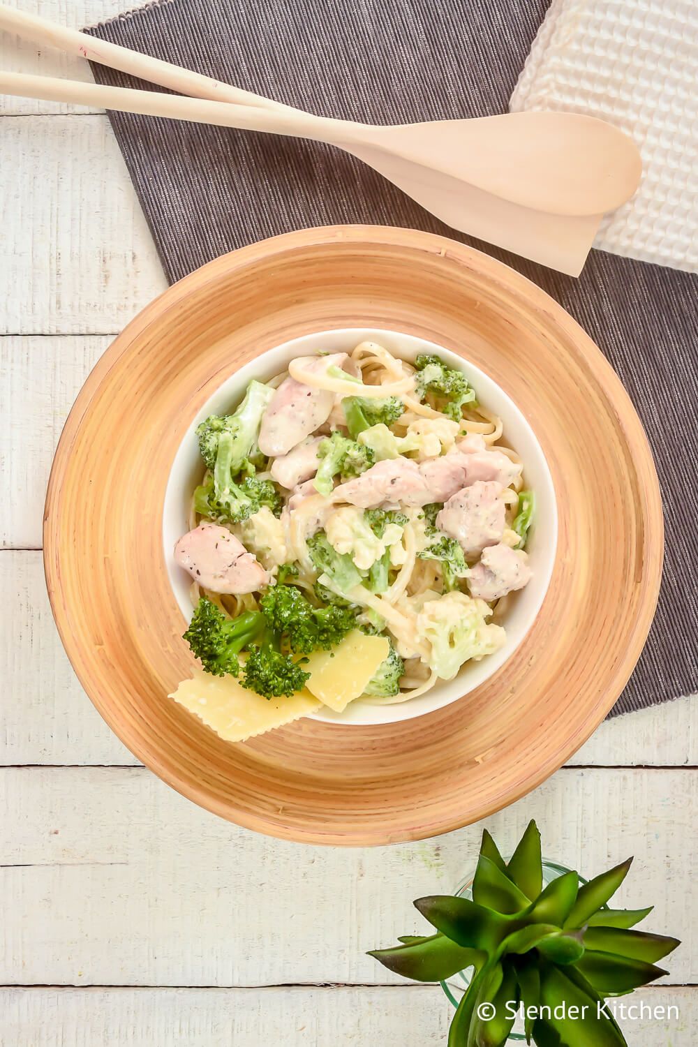 Lightened up chicken broccoli alfredo in a bowl with a wooden plate and wooden utensils.