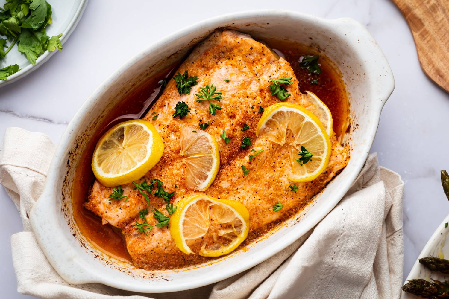 Lemon pepper salmon with lemon slices, butter, and lemon pepper seasoning in a white baking dish.