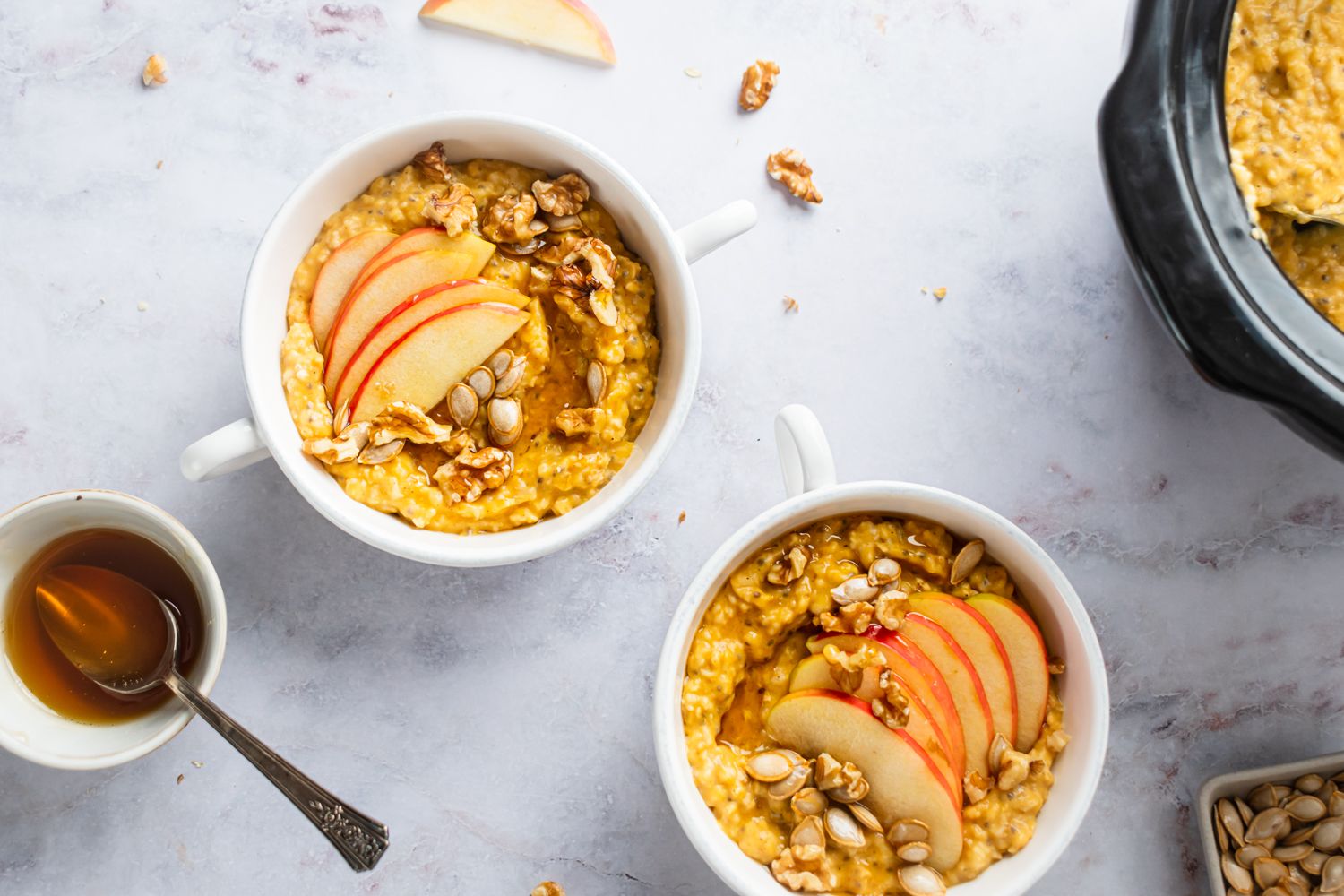 Crockpot steel cut pumpkin oatmeal with sliced apples and pepitas in two mugs.