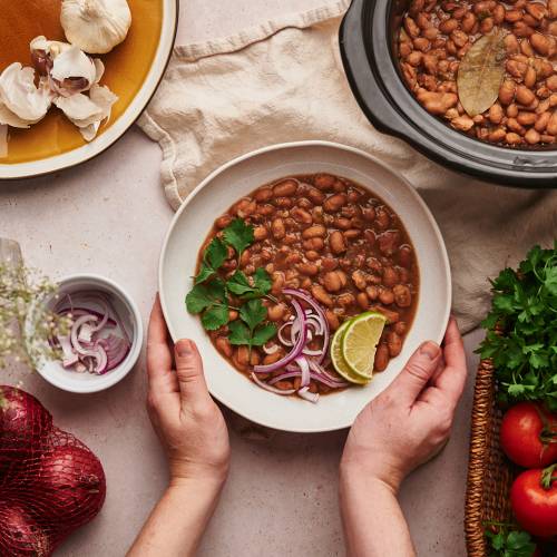 Crockpot pinto beans in a bowl with red onions, cilantro, lime slices, and other vegetables.