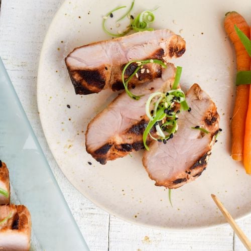 Asian grilled pork tenderloin on a plate with carrots, green onions, and chopsticks.