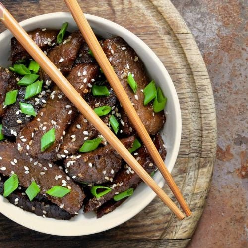 Mongolian Beef in a bowl with chopsticks with scallions and sesame seeds.
