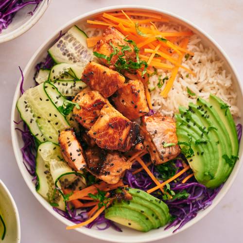 Salmon rice bowl with salmon pieces that are crispy served with rice, purple cabbage, edamame, carrots, and cucumber.