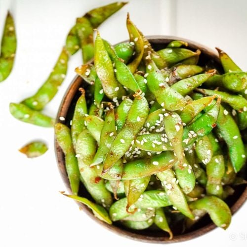 Stir fried edamame in a wooden bowl served with soy sauce and sesame seeds.