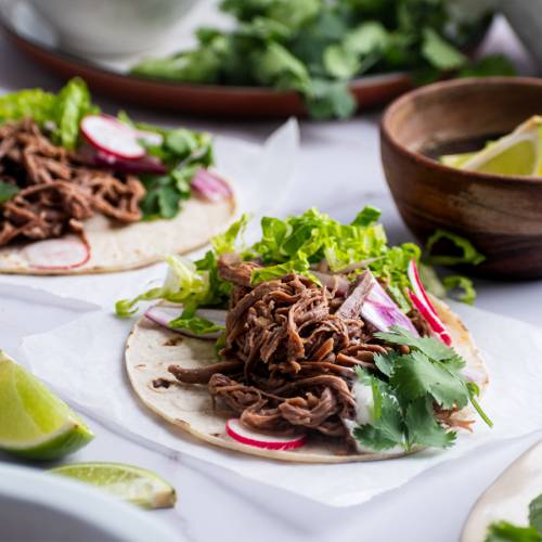 Slow cooker tri tip tacos on white corn tortillas with lettuce, cilantro, and radishes.