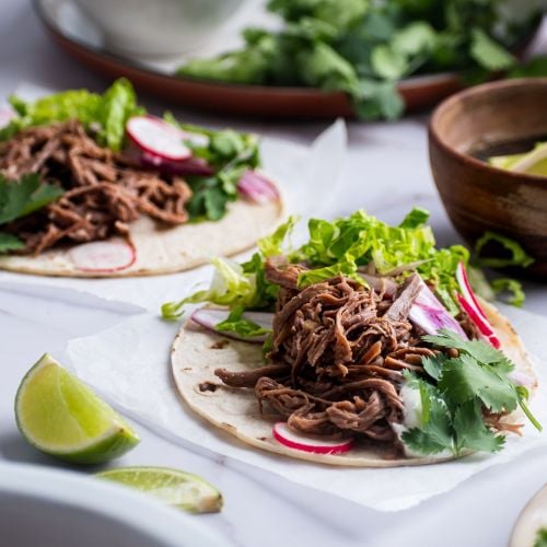 Slow cooker tri tip tacos on white corn tortillas with lettuce, cilantro, and radishes.