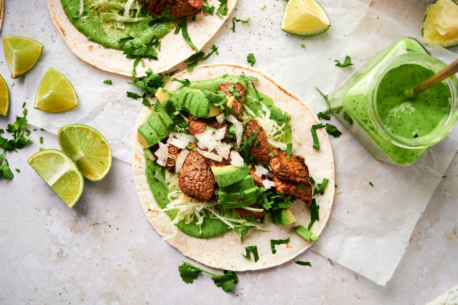 Carne asada tacos with tender steak, cilantro, onion, lime juice, avocado, and green salsa in two corn tortillas.