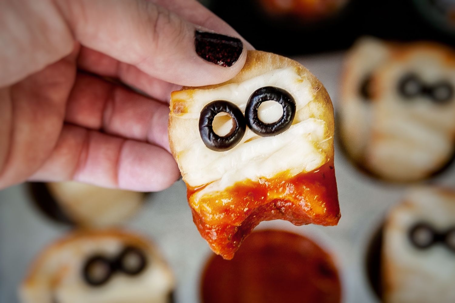 Mummy pizza flatbread being held above a muffin tray with marinara sauce for dipping.