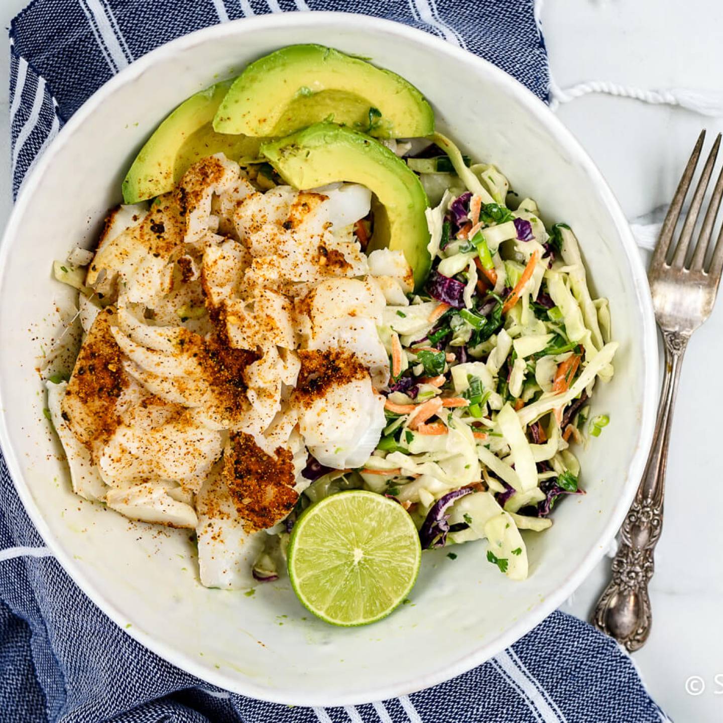 Ancho fish taco bowl with lime slaw and sliced avocado with a blue napkin.