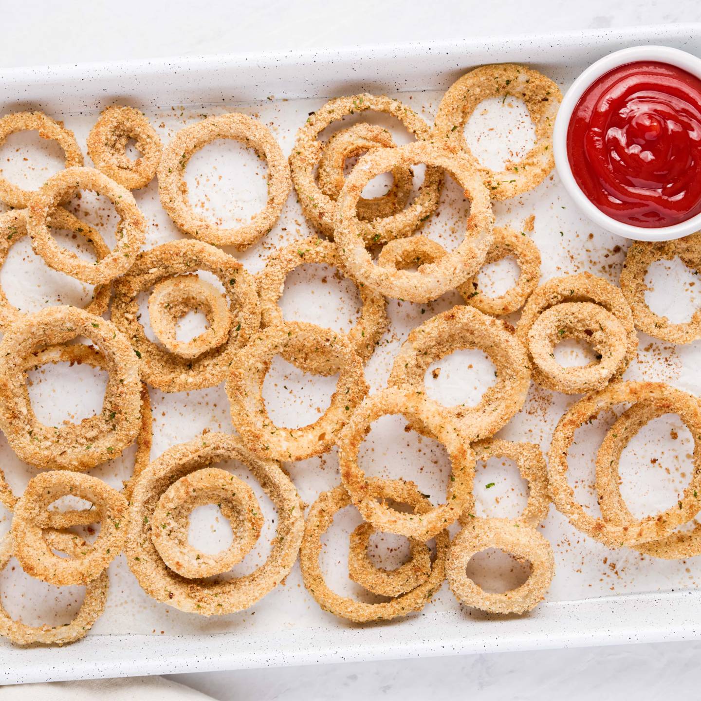 Baked onion rings with a golden brown breadcrumb coating on a baking sheet with ketchup on the side.