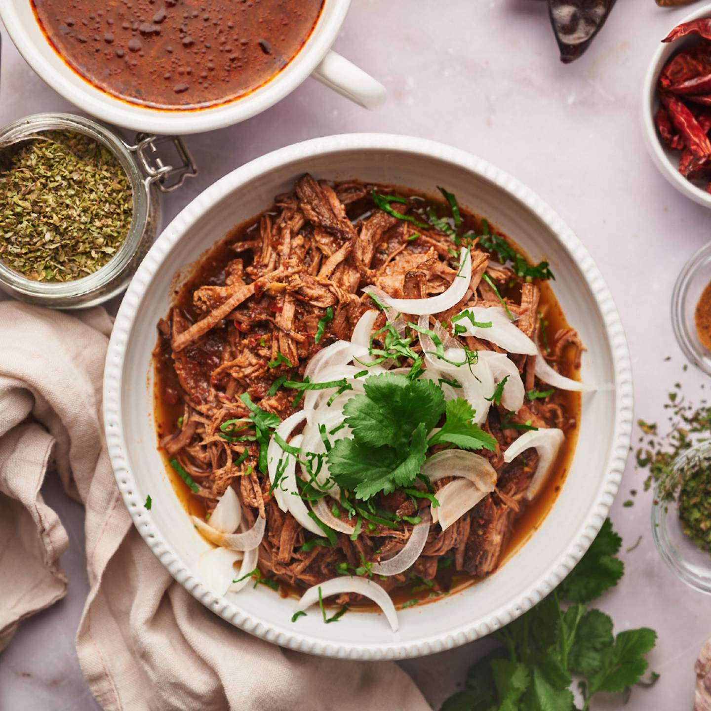 Birria de res with a red chile sauce served in a bowl with corn tortillas, sliced onions, cilantro, and consome.