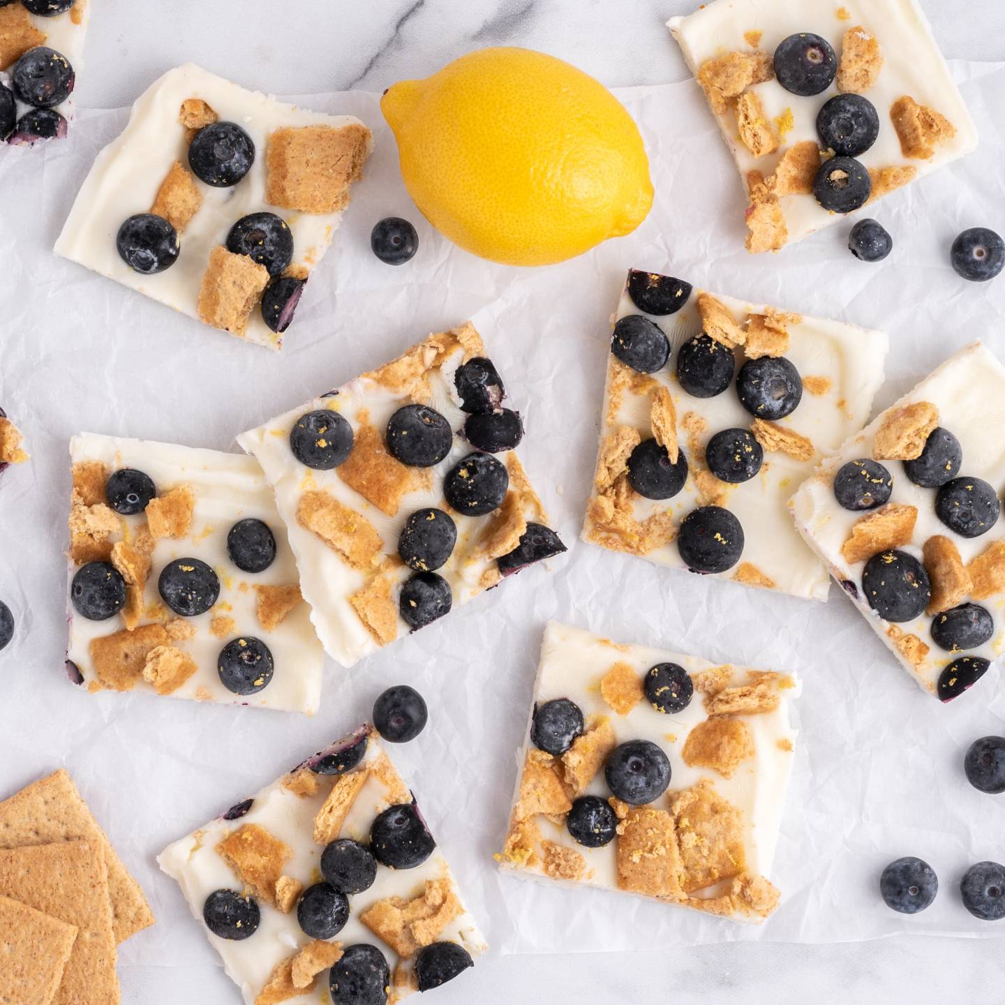 Blueberry lemon frozen yogurt bark with graham crackers on a white board with fresh lemon and blueberries.