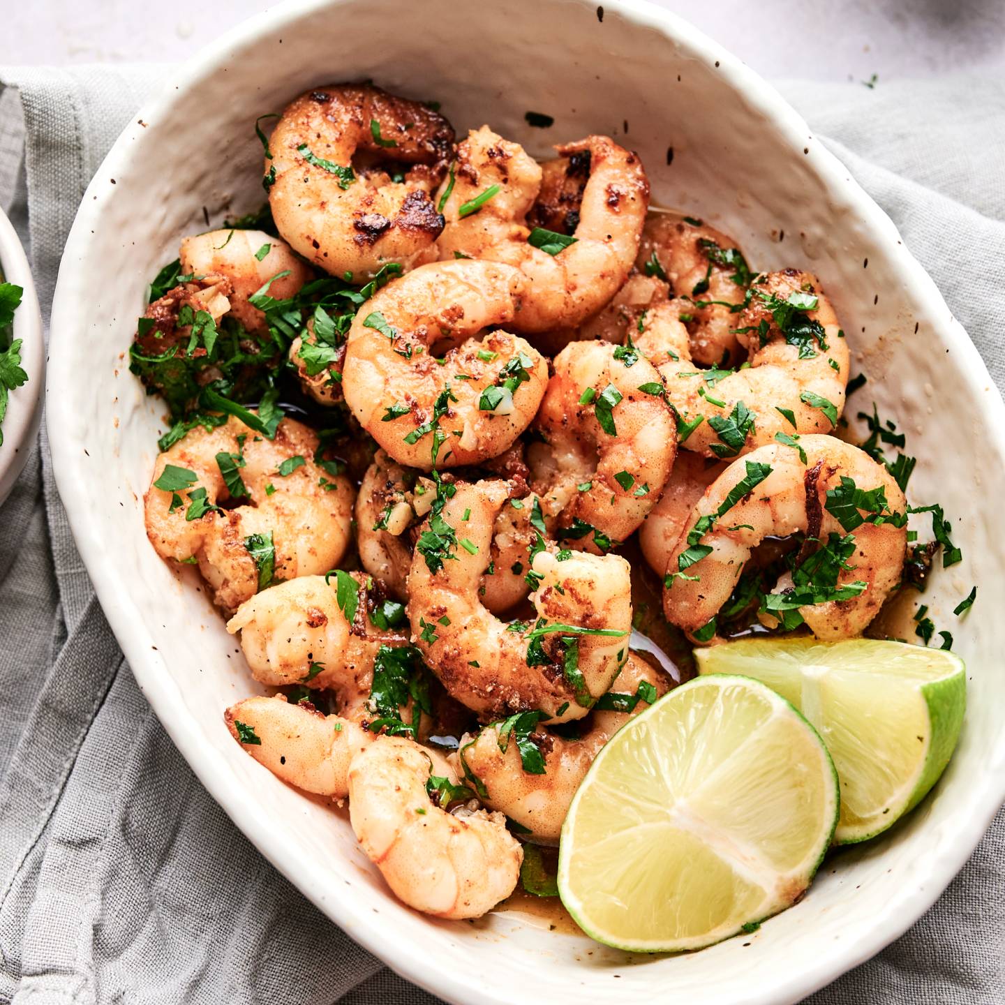 Camarones al ajillo in a white dish with butter garlic sauce, parsley, chile peppers, and lime wedges. 