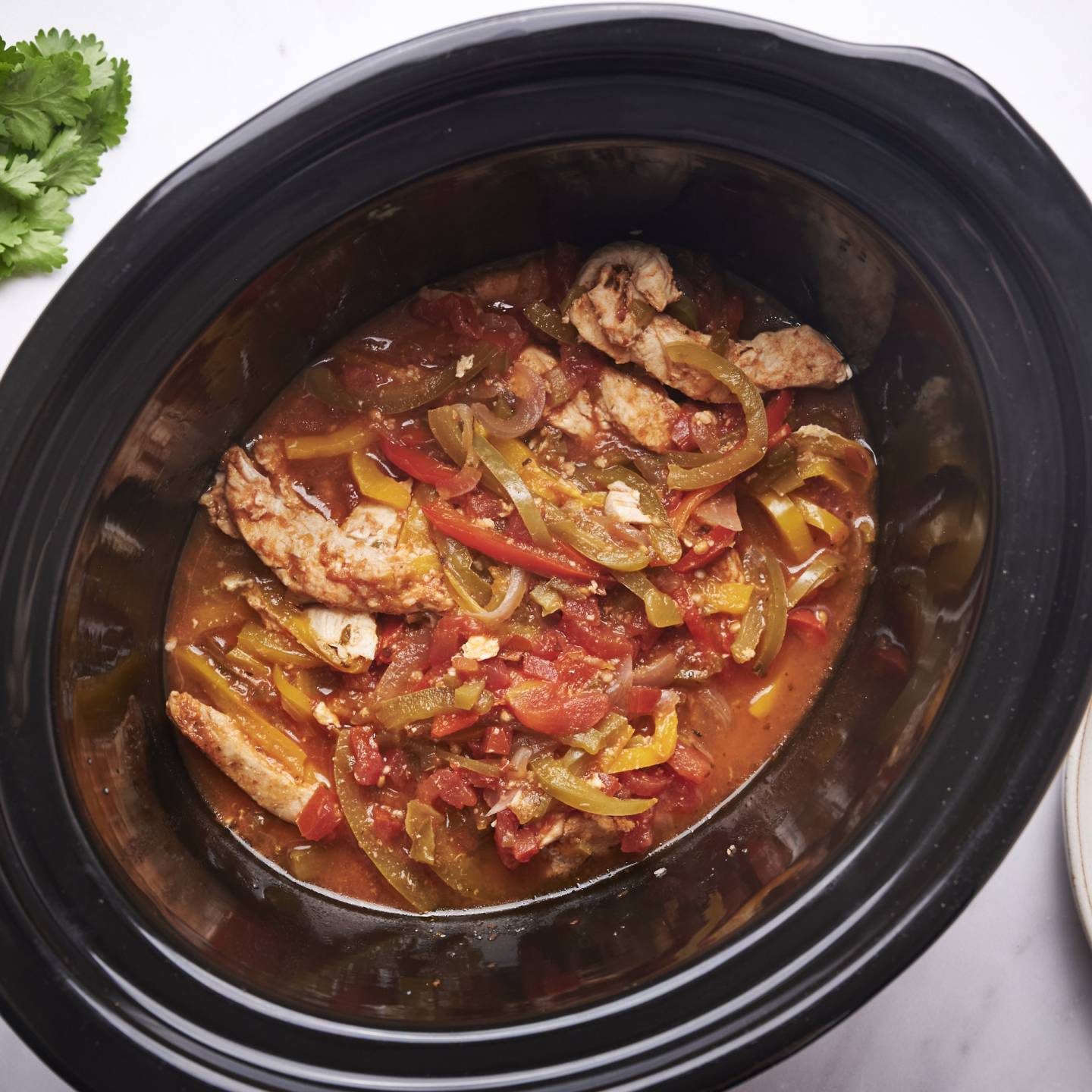 Slow cooker chicken fajitas with bell peppers, onions, sliced chicken breast, and tomatoes in a black slow crockpot with tortillas on the side.