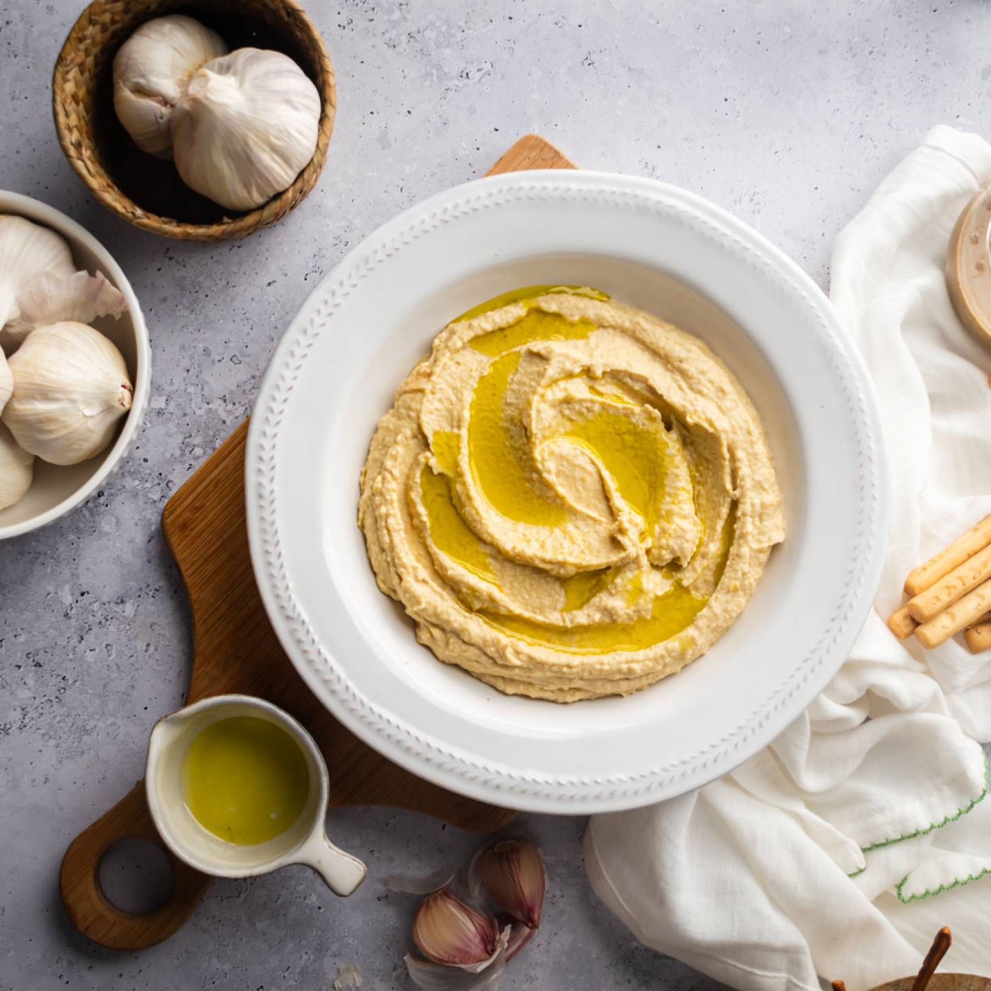 Garlic hummus swirled with olive oil with garlic cloves and breadsticks on the side.