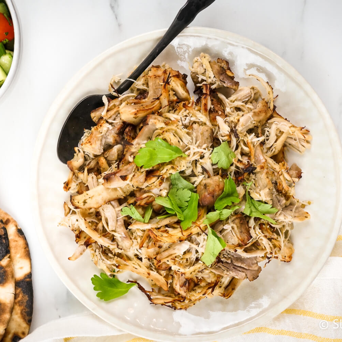 Greek chicken shredded on a plate with oregano, parsley, lemon, pita bread, and cucumber tomato salad.