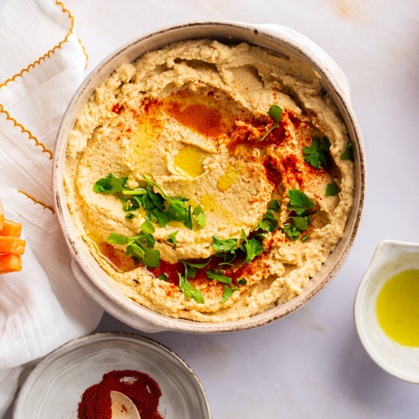 Hummus made with chickpeas in a bowl with olive oil, parsley, and paprika with carrots and pretzels on the side.