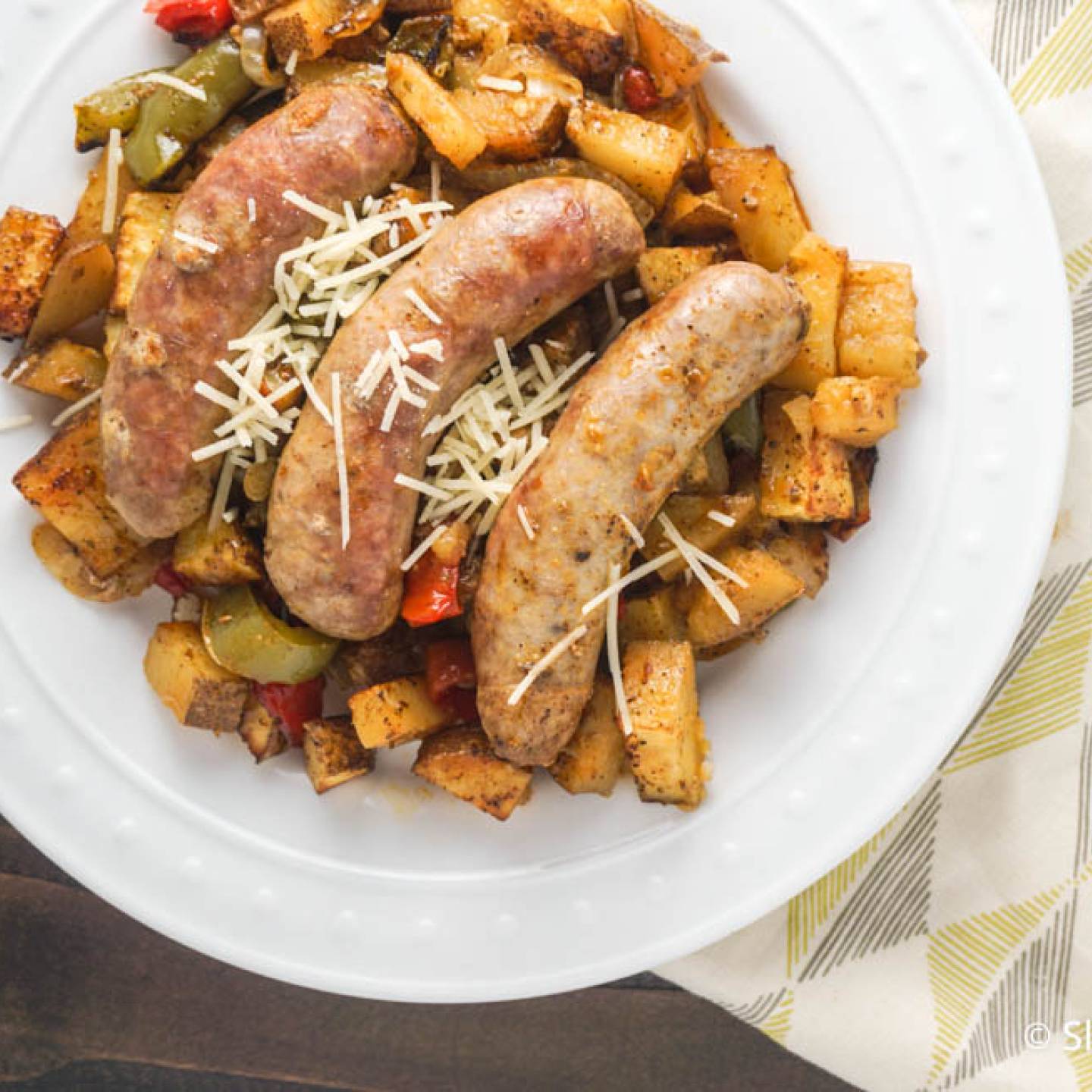 Roasted sausages with potatoes, peppers, and onions on a plate with Parmesan cheese.