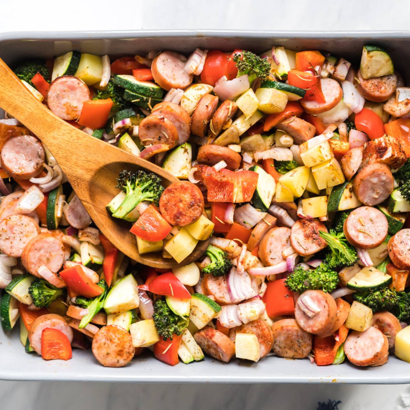 One pan sausage, potato, and veggie in a baking dish with a wooden spoon.