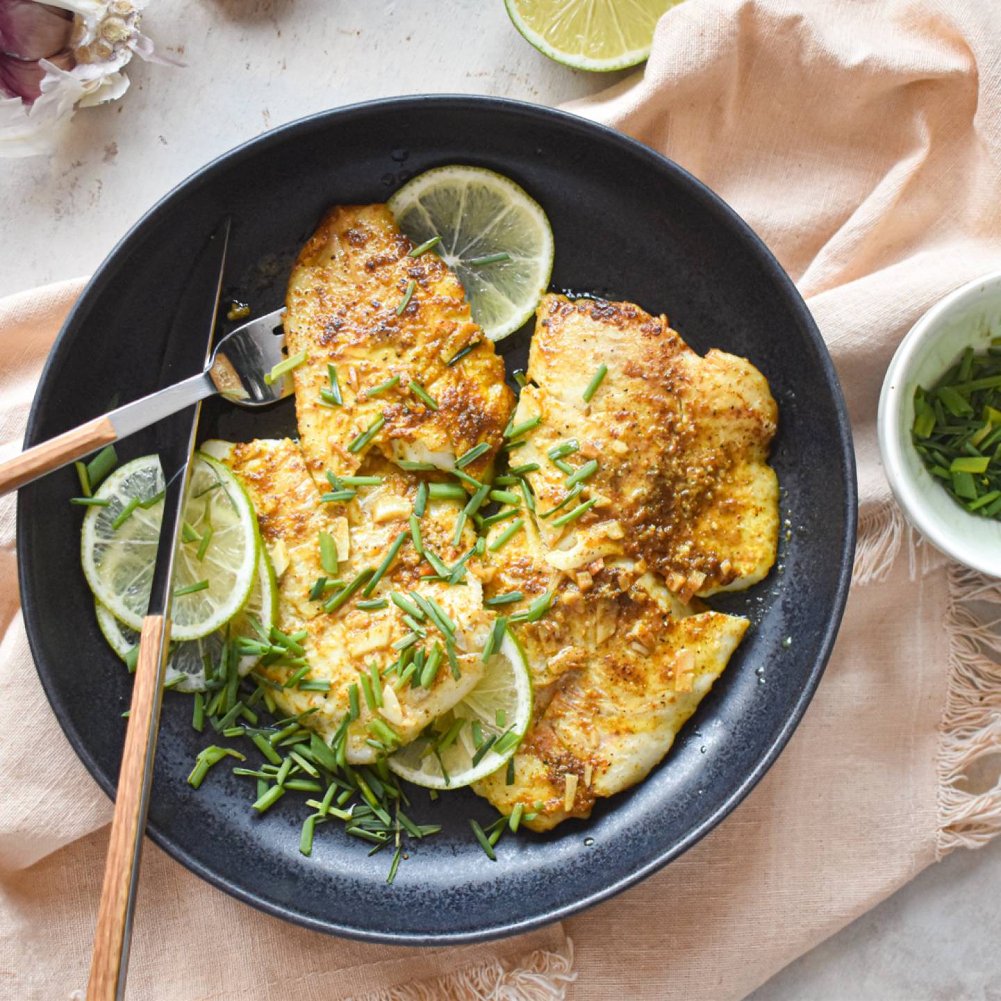 Pan fried garlic lime tilapia on a plate with fresh limes, green onions, and garlic.