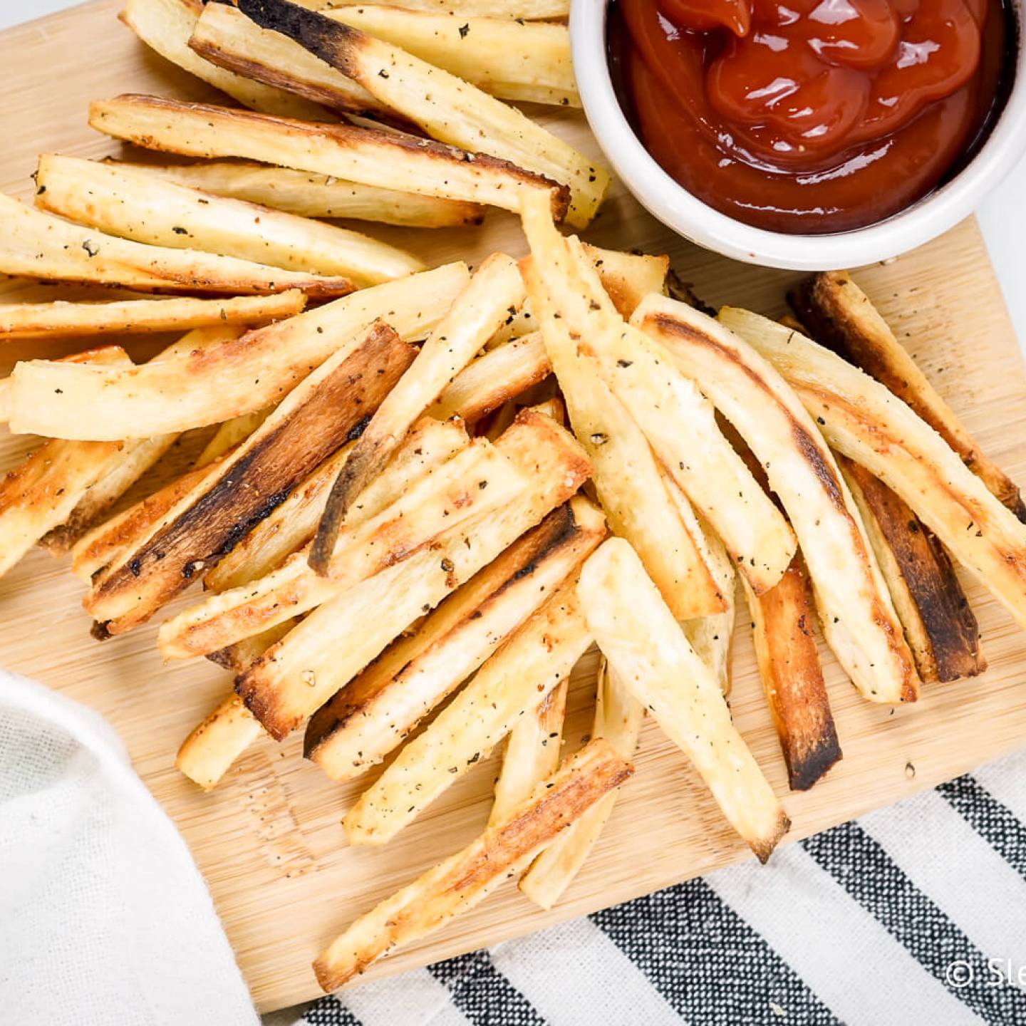 Parsnip fries cut into match sticks with spices on a wooden cutting board with kethcup.