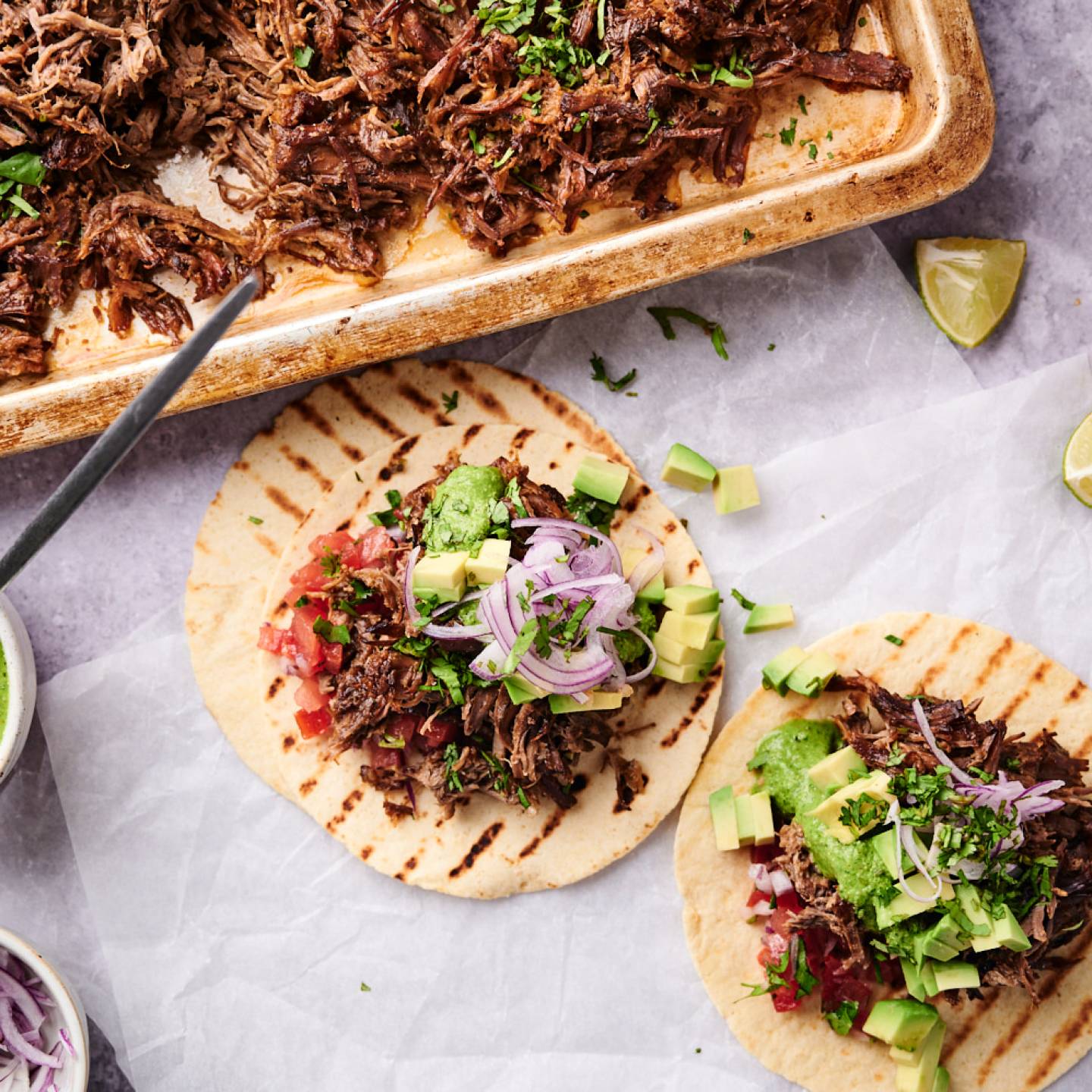 Crockpot carne asada served on tortillas with avocado, pico de gallo, red onion, and lime wedges.