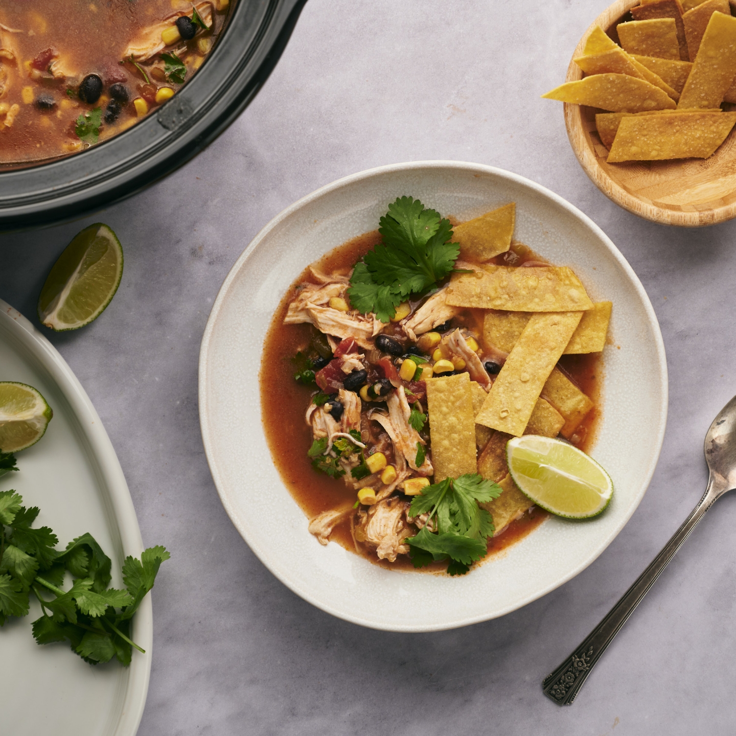 Slow cooker chicken tortilla soup with shredded chicken, beans, corn, cilantro, crispy tortilla strips, and lime wedges.