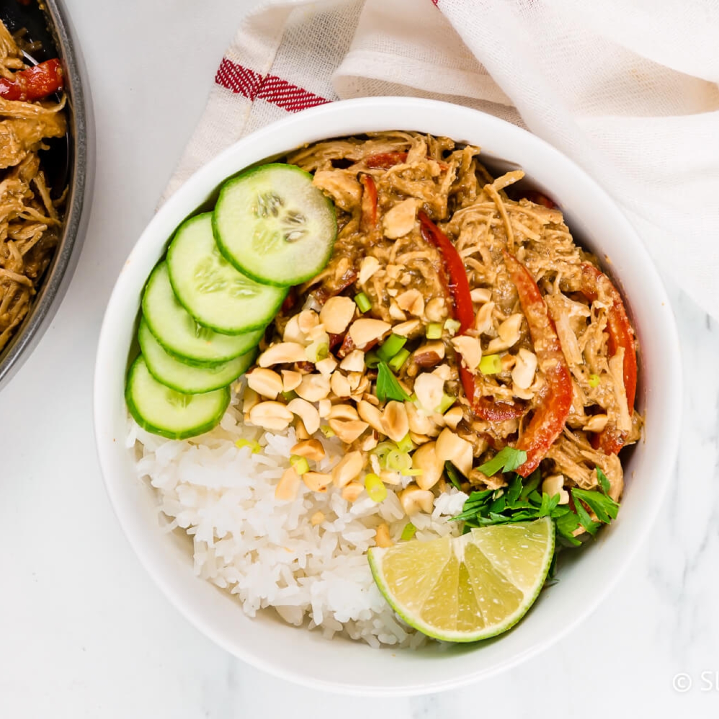 Slow Cooker Thai Peanut Chicken in a bowl with rice, cucumbers, chopped peanuts, and lime. 