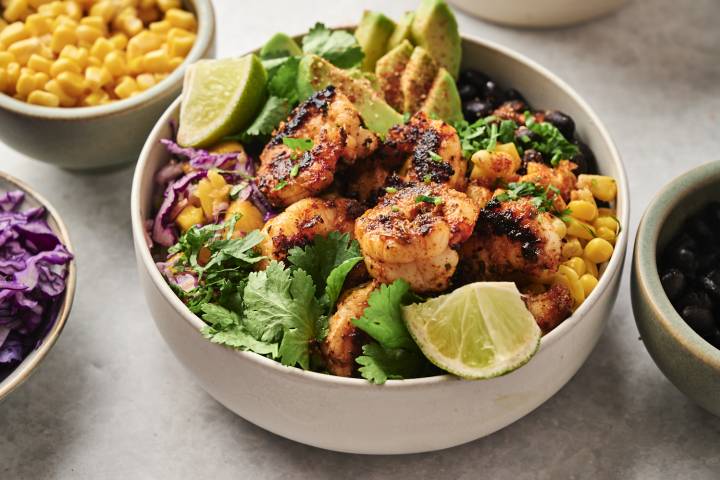 Blackened shrimp bowl topped with grilled shrimp, avocado slices, lime wedges, cilantro, and surrounded by corn, purple cabbage, and black beans.