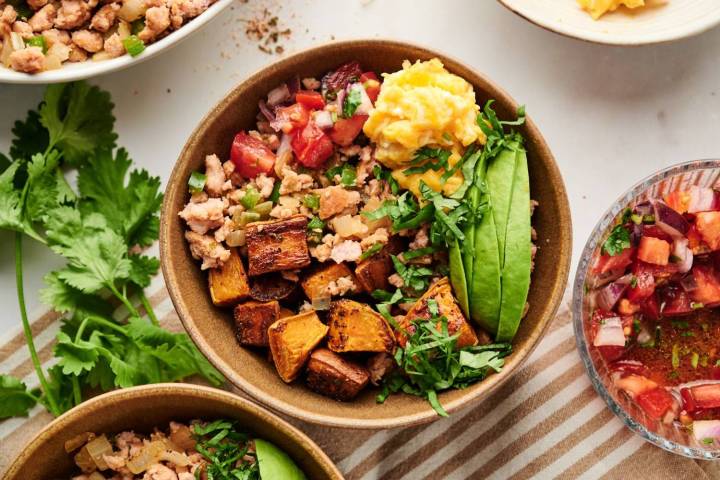 A vibrant breakfast burrito bowl filled with scrambled eggs, roasted sweet potatoes, seasoned ground meat, fresh avocado slices, and a sprinkle of chopped cilantro.