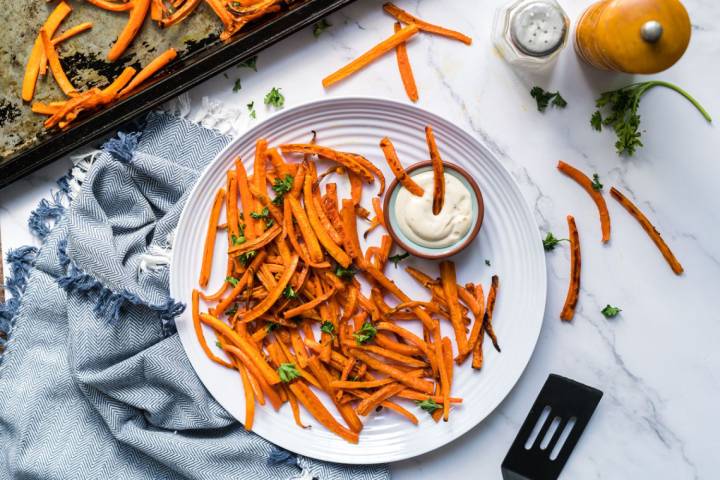 Crispy baked carrot fries garnished with fresh parsley on a white plate, served with a small bowl of creamy dipping sauce. The plate is placed on a marble countertop, surrounded by a baking tray, a blue fringed napkin, and scattered carrot fries and parsley.