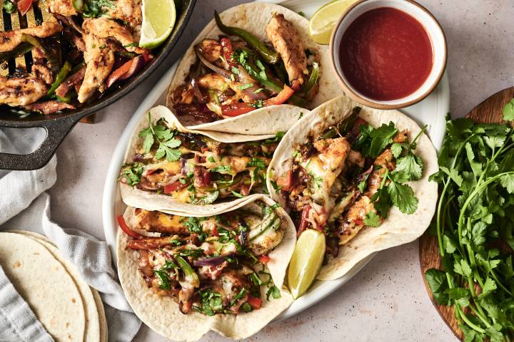 A plate of chicken alambre tacos topped with cilantro, melted cheese, and lime wedges, with a side of salsa, accompanied by a skillet of sautéed chicken and vegetables.