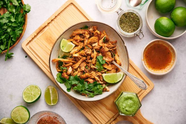 Chicken carnitas in a bowl sitting on a wooden cutting board with ingredients around it. 