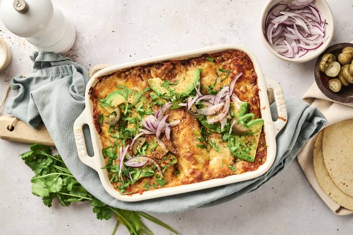 A close-up of cheesy chicken enchiladas being served from a casserole dish, topped with avocado, red onion, jalapeños, and fresh cilantro.